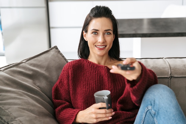 Image of an amazing woman indoors in home on sofa watch TV drinking coffee.