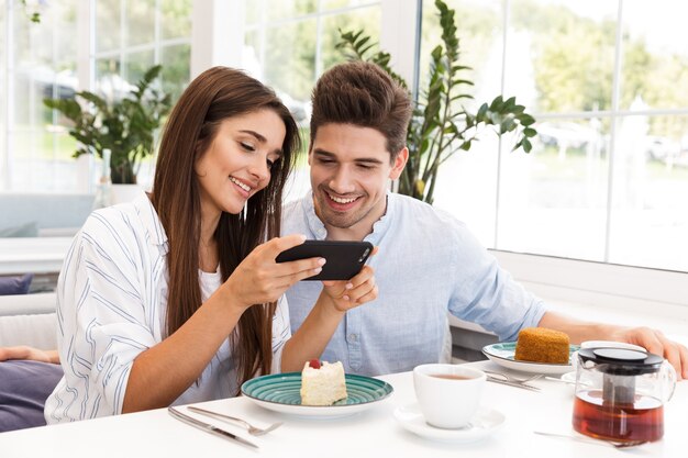 Image of amazing young loving couple sitting in cafe eat desserts and drinking tea talking with each other holding mobile phone.