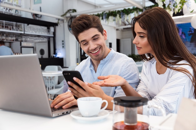 Image of amazing young loving couple sitting in cafe using laptop computer and mobile phone.