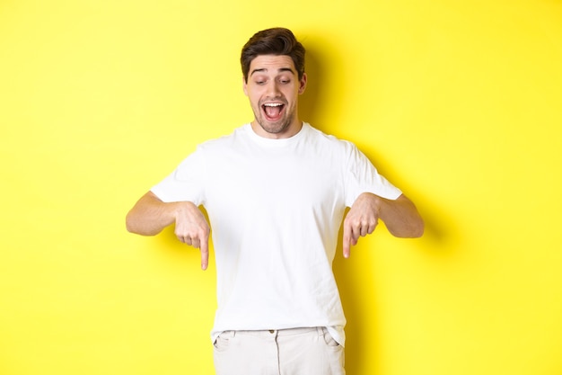 Image of amused handsome guy in white t-shirt, looking and pointing fingers down, standing over yellow wall