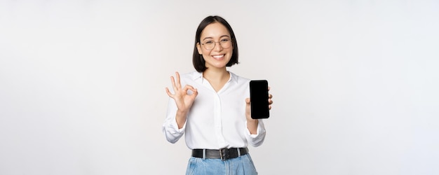 Image of asian businesswoman showing smartphone screen app interface and ok sign recommending application on mobile phone standing over white background