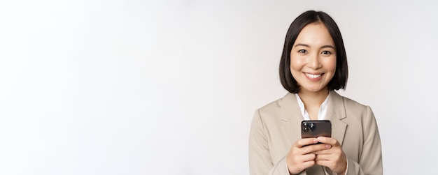 Image of asian businesswoman in suit holding mobile phone using smartphone app smiling at camera whi