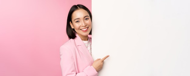 Image of asian female entrepreneur in suit pointing finger at board showing smth on white wall demonstrating chart or information standing over pink studio background