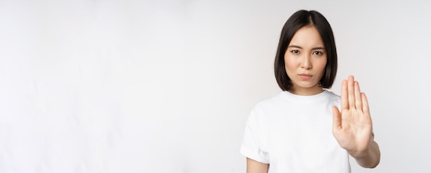 중지 금지 smth를 보여주는 아시아 소녀의 이미지는 흰색 배경 위에 티셔츠를 입고 서 있는 금기 제스처를 보여주기 위해 한쪽 팔을 뻗습니다.