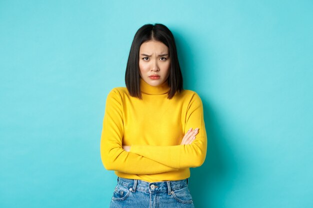 Image of asian girlfriend feeling offended, cross arms on chest and sulking, staring at camera insulted with something, standing over blue background.