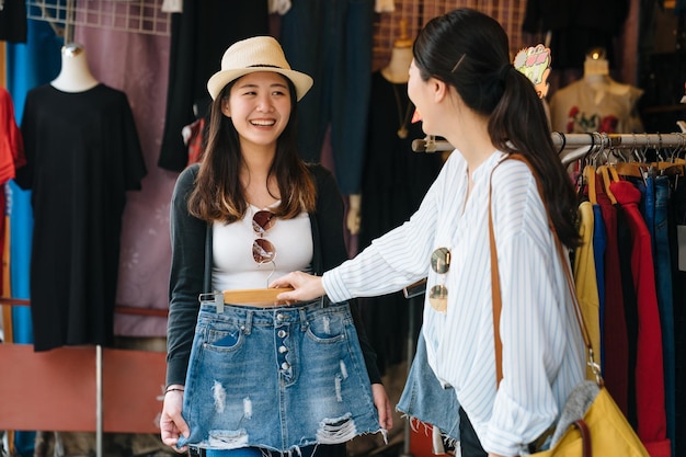 Image of asian japanese charming girl trying on fashionable jeans while friend giving advising and holding clothes rack. two women friends having fun and laughing during shopping outdoor local market