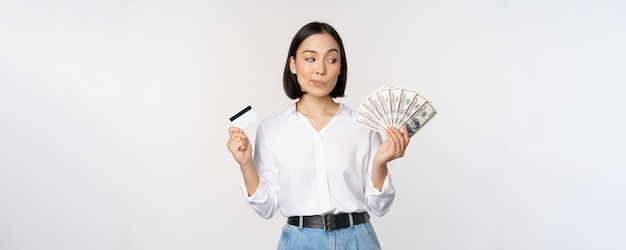 Image of asian woman looking at money dollars holding credit card in another hand thinking standing