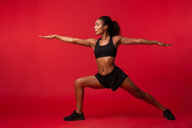 Image of athletic african american woman in black sportswear stretching her body, isolated over red wall