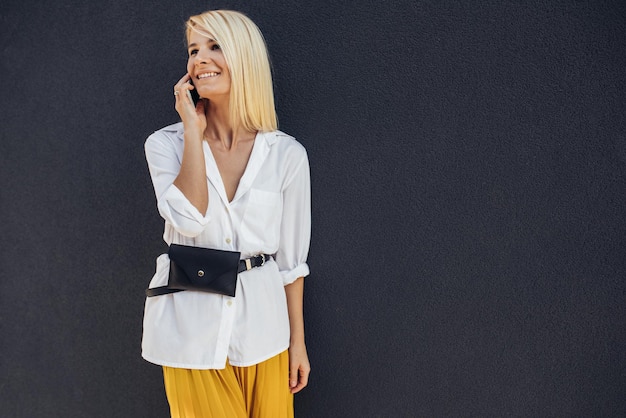 Image of attractive blonde business woman standing in a gray building wall while talking on her mobile phone looking away Beautiful Caucasian female dressed in white shirt an yellow skirt Copy space