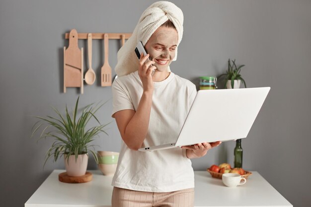 Image of attractive busy woman wearing white Tshirt and wrapped in towel with beauty treatment facial mask using her laptop and cell phone for freelance work expressing positive emotions