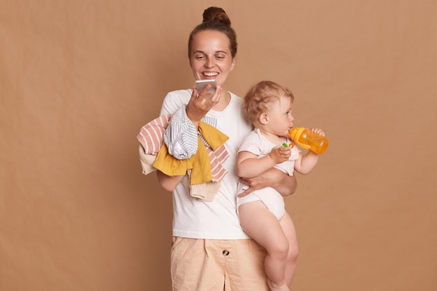 Image of attractive happy joyful woman with bun hairstyle wearing white t shirt standing with her baby daughter and using cell phone for recording voice message isolated over brown background