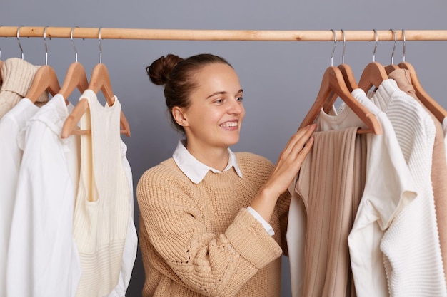 베이지색 스웨터를 입고 쇼핑몰에서 포즈를 취하고 있는 매력적인 긍정적인 여성의 이미지는 선반에 있는 옷장에 매달린 옷을 입고 회색 벽에 서서 쇼핑을 즐기는 새로운 옷을 선택합니다.