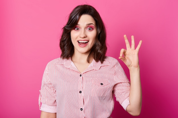 Image of attractive stylish brunette girl in striped shirt looking at camera with opened mouth and showing ok gesture