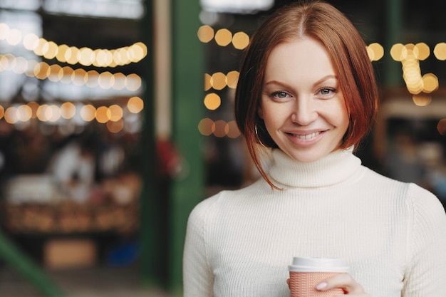 Image of attractive young woman with brown hair gentle smile wears white turtleneck sweater holds paper cup of coffee has pleased expression poses over blurred background with copy space