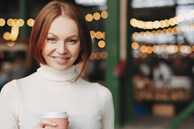Image of attractive young woman with brown hair gentle smile wears white turtleneck sweater holds paper cup of coffee has pleased expression poses over blurred background with copy space