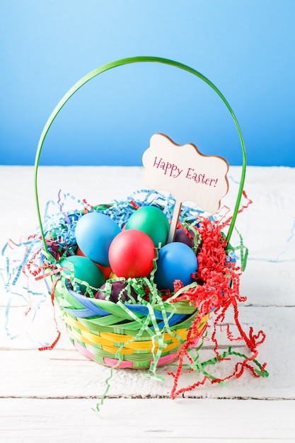 Image of basket with colorful eggs on empty blue wall on wooden table with wish for happy Easter