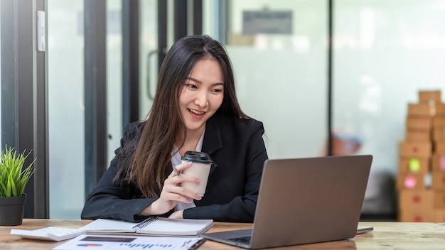 Image of a beautiful Asian businesswoman happy a successful work holding a coffee cup  with a laptop and document at the office.