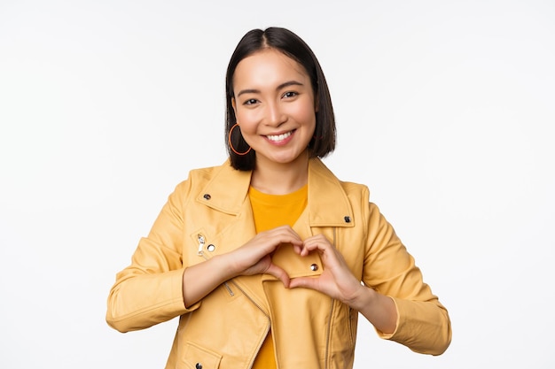 Image of beautiful asian girl showing heart love sign express care and romantic feelings standing over white studio background