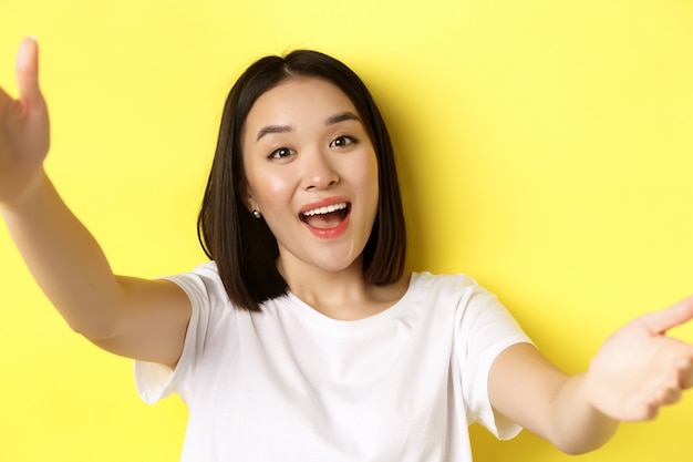 Image of beautiful asian woman holding smartphone camera and smiling, taking selfie or video chat, standing over yellow background.