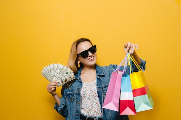 Image of a beautiful blonde girl smiling with sunglasses, holding shopping bags  and money over yellow wall
