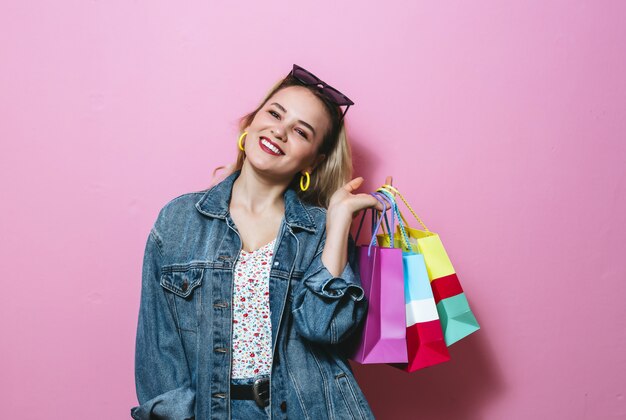 Image of a beautiful blonde girl smiling with sunglasses, holding shopping bags  over pink wall