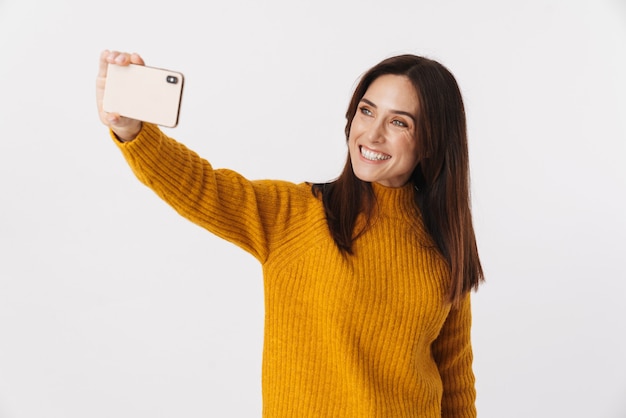 Image of beautiful brunette adult woman wearing sweater smiling and taking selfie photo on cellphone isolated on white