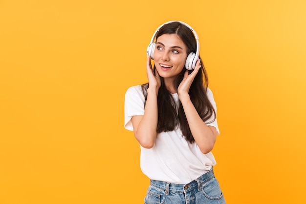 Image of beautiful brunette woman with long hair smiling while listening to music with headphones isolated over yellow wall