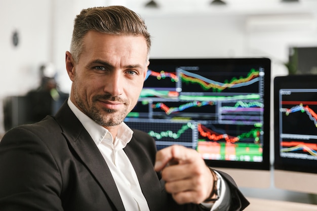 Image of beautiful businessman 30s wearing suit working in office on computer with graphics and charts at screen