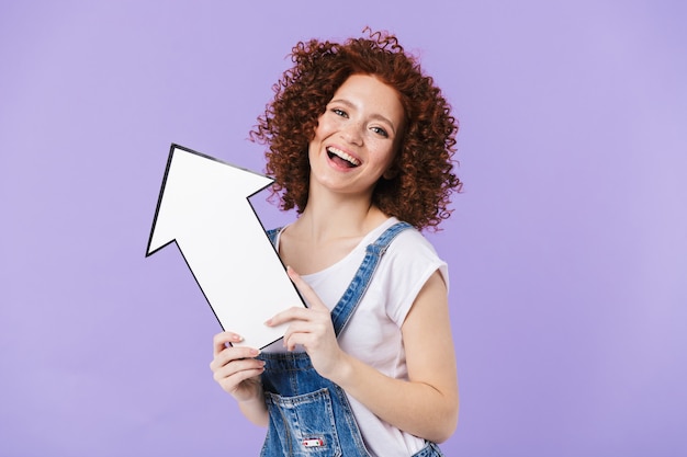 Image of a beautiful curly happy redhead girl posing isolated over purple wall holding arrow.