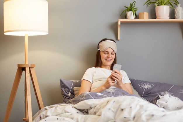 Image of beautiful dark haired caucasian woman wearing blindfold and sleepwear holding mobile phone sitting in bed under blanket in home interior expressing positive emotions