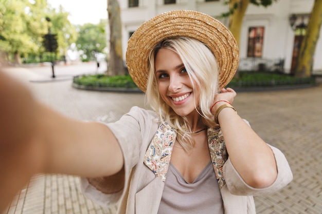 Image of beautiful lady wearing suit and straw hat smiling, while taking selfie photo outdoor in downtown