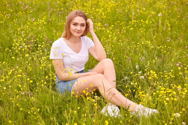 Image of beautiful pretty happy smiling girl sitting on green summer meadow and looking directly 