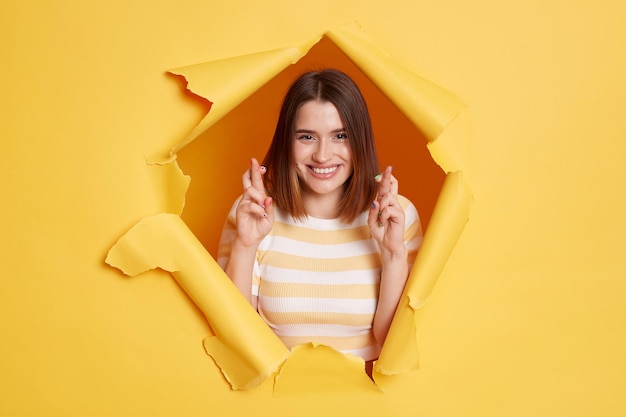 Immagine di bella donna attraente sorridente che indossa una t-shirt a righe guardando attraverso carta gialla strappata incrociando le dita sorridendo alla fotocamera che esprime il desiderio