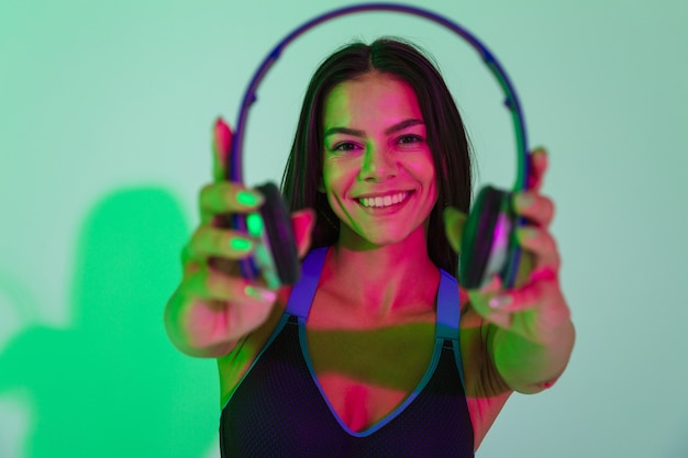 Image of beautiful smiling optimistic young strong fitness woman isolated with led flash lights give you a headphones.