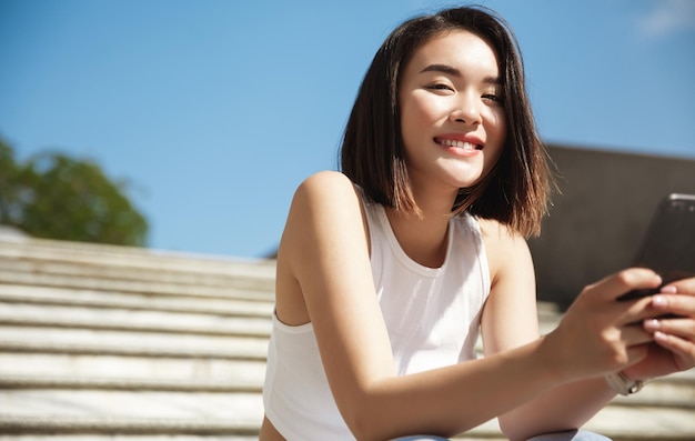 Immagine di una bella donna alla moda seduta sulle scale della strada in un giorno d'estate e con in mano un telefono cellulare ragazza asiatica invia un messaggio di testo sullo smartphone e sorride alla telecamera