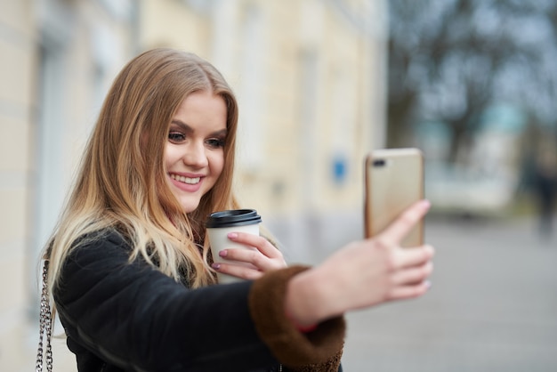 Image of beautiful woman holding takeaway coffee in paper cup and taking selfie while walking through city street