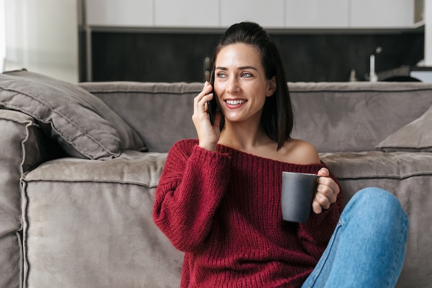 コーヒーを飲みながら携帯電話で話しているソファの近くの家の屋内で美しい女性の画像。