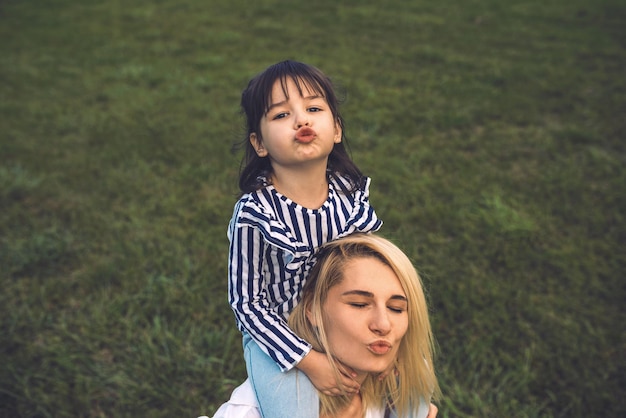 Image of beautiful woman playing and blowing kiss with her cute little girl on green grass outdoor Loving pretty mother and daughter spend time together in the park Mom and kid have funHappy family