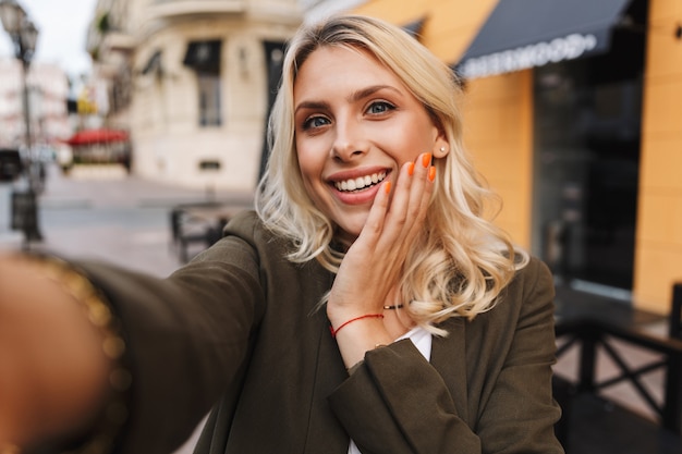 Image of beautiful woman smiling and taking selfie photo, while walking through city street