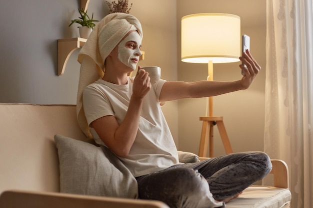 Image of beautiful woman in white Tshirt and pajama pants wrapped in white towel sitting on sofa with cosmetic mask doing cosmetology procedures drinking coffee and making selfie on smart phone