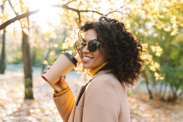 Image of a beautiful young african woman walking outdoors in a spring park drinking coffee.