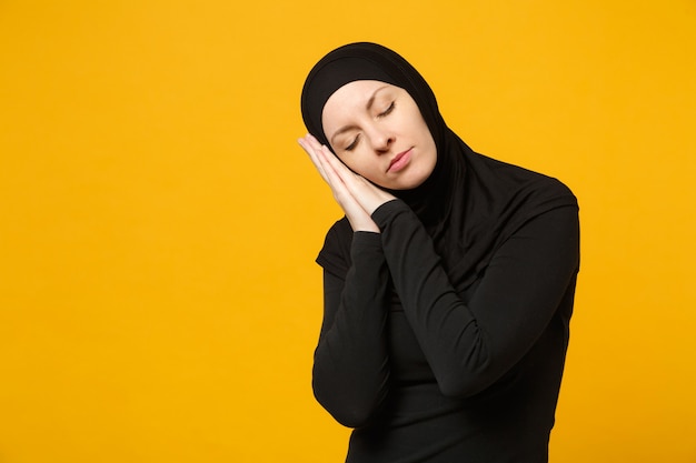 Hijab 검은 옷에 아름 다운 젊은 아라비아 이슬람 여자의 이미지 노란색 벽에 고립 된 뺨 아래 접힌 손으로 잠. 사람들이 종교 이슬람 라이프 스타일 개념 복사 공간을 모의