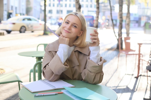 Изображение красивой молодой блондинки, пьющей кофе в открытом кафе, работающей в университете