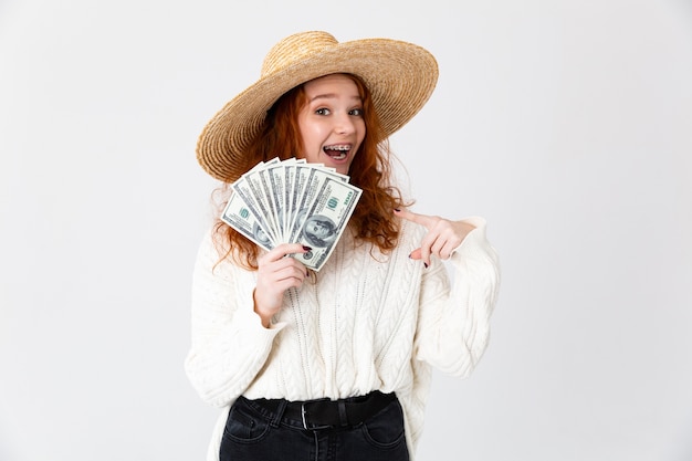 Image of a beautiful young cute girl redhead posing isolated over white wall background wearing hat holding money.