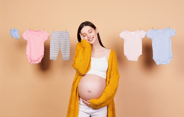 Immagine di una bellissima giovane donna emotiva incinta carina in posa isolata su uno sfondo rosa circondata da vestiti per bambini su una corda, che si prepara al parto, l'aspetto di un bambino.