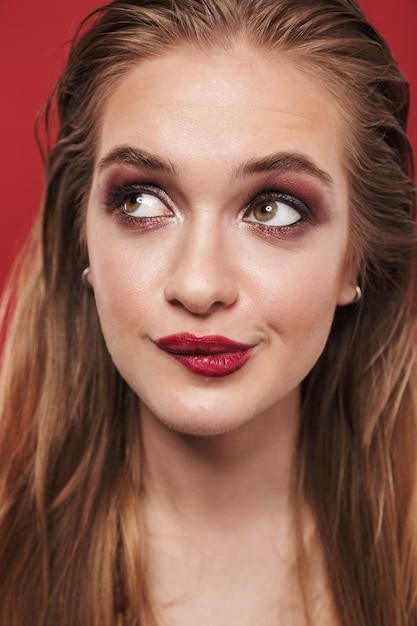 Image of a beautiful young dreaming amazing woman with bright makeup red lipstick posing over red wall.