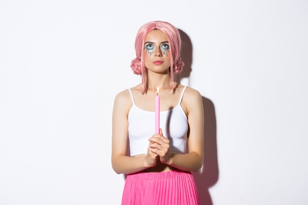 Image of beautiful young party girl in pink wig, holding candle to cast a spell on halloween , standing over white background serious.