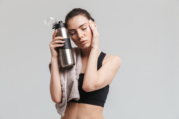 Изображение красивой молодой усталой женщины фитнеса спорта представляя с полотенцем и бутылкой с водой, изолированной над серой стеной.