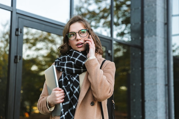 Image of a beautiful young woman outdoors walking by street holding laptop computer talking by mobile phone.