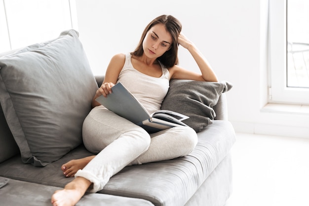 Image of beautiful young woman sitting on sofa indoors at home reading book.